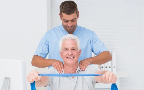 A man holding a resistance band and stretching while a physical therapist adjusts his shoulders